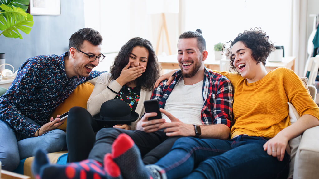 4 amis assis dans le canapé de leur coliving qui rigolent en regardant quelque chose sur un smartphone