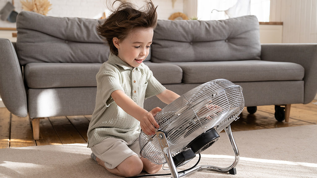 Petit garçon qui se rafraichît avec un ventilateur dans son salon